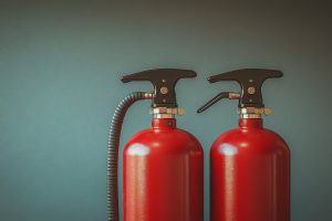 Two red fire extinguishers are displayed prominently against a teal background, showcasing their important role in fire safety at indoor locations, emphasizing readiness and functionality.